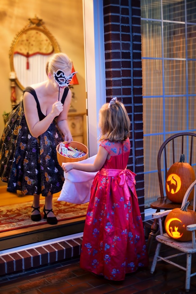 Woman with a masquerade mask giving candy to a trick or treater dressed as a princess. 